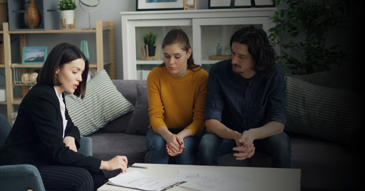 A real estate agent sits at a drawing room table with a curious young couple, going over loan options for their dream home.