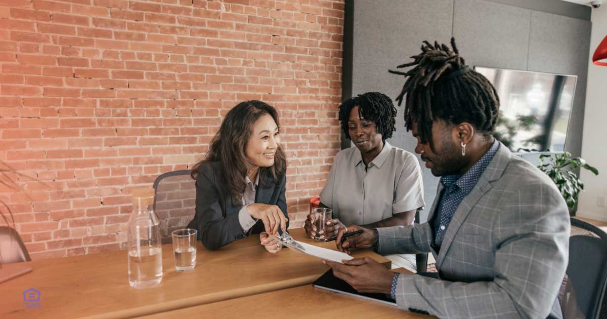 A Black couple sits at a dining table with a mortgage officer, reviewing home equity options in their living room. The advisor points to a document, offering guidance on tapping into property value for financial goals.